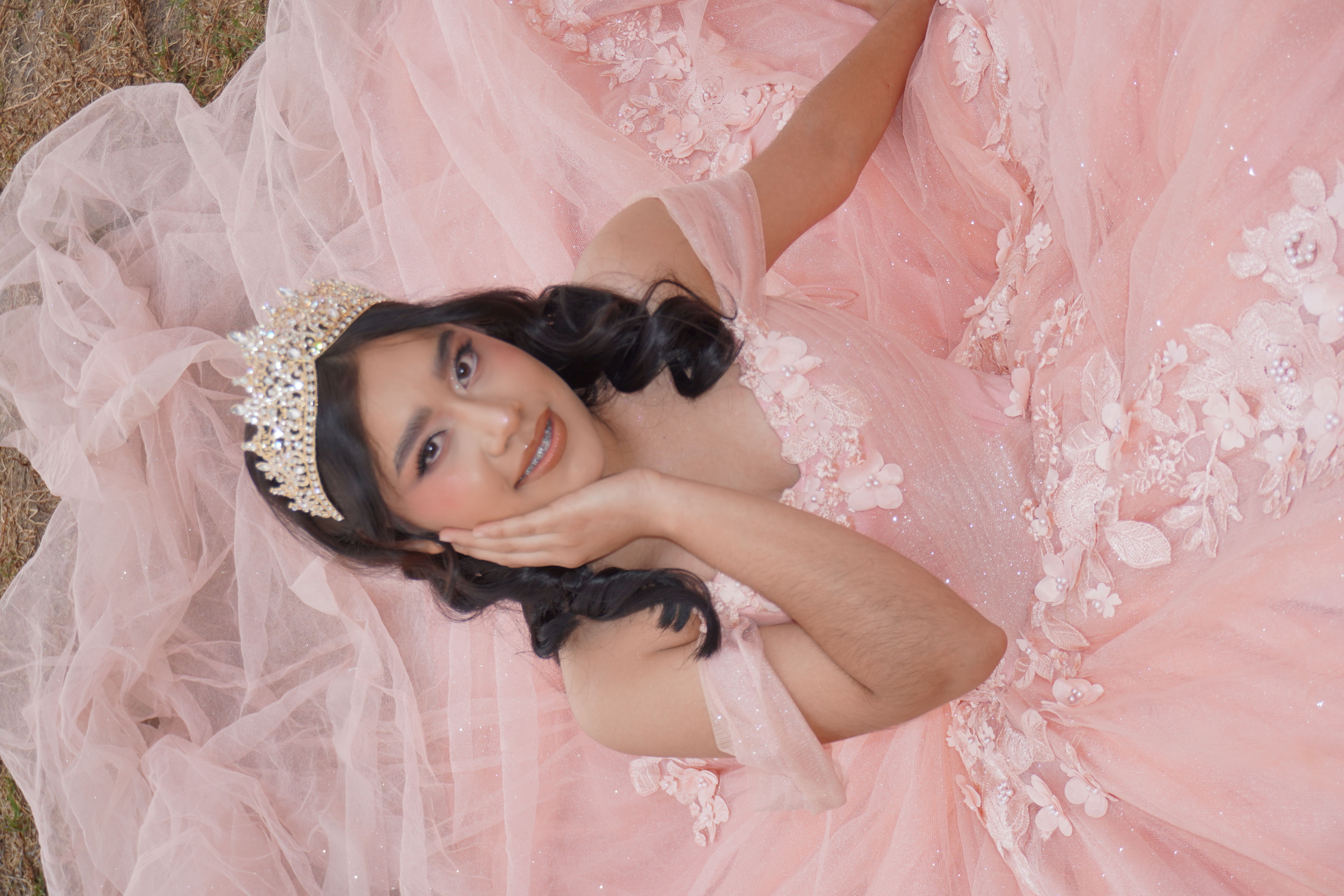 Quinceañera de rosa recostada sobre tul con corona dorada y sonrisa