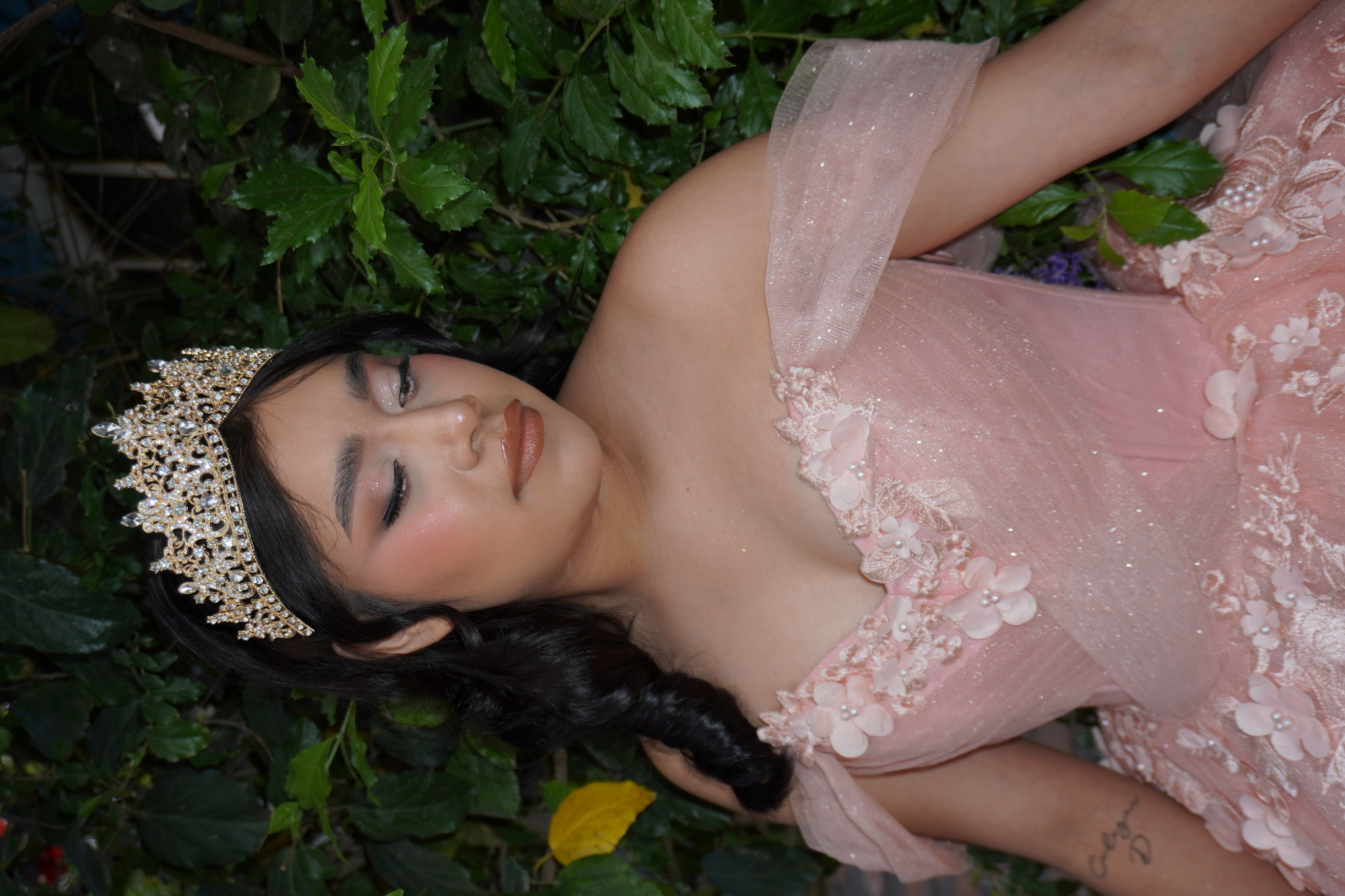Quinceañera de rosa con corona dorada y mirada baja frente a un seto verde